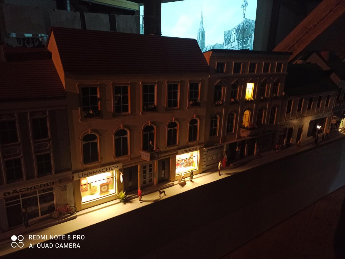 Minmiatura miasteczka nocą z podświetlonymi witrynami sklepów: zegarmistrz, piekarz i inne. W tle czarno-biała panorama.